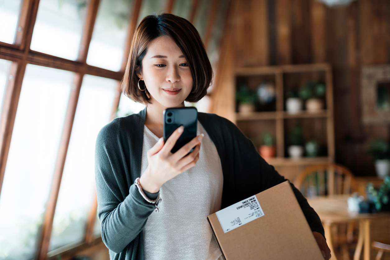 Woman looking at smartphone holding a package to delivery for her eCommerce store - Shift4Shop